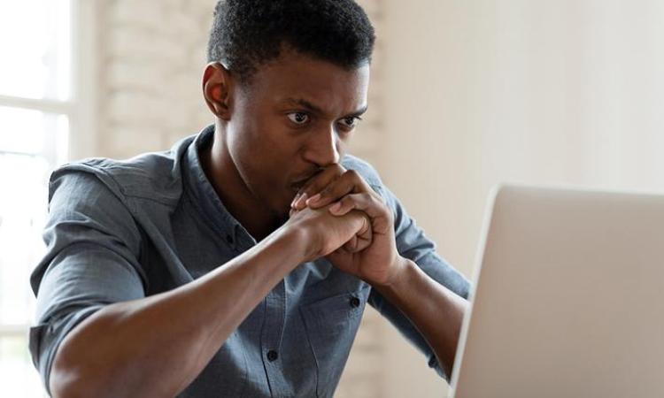 A young Black man in front of a computer, thinking, with his hands covering his mouth.