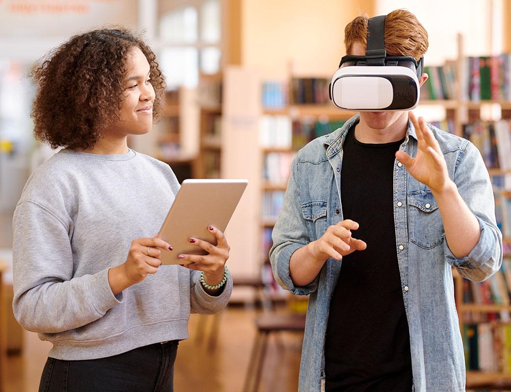 A young Black woman with a notebook smiles while watching a young white man wearing VR glasses.